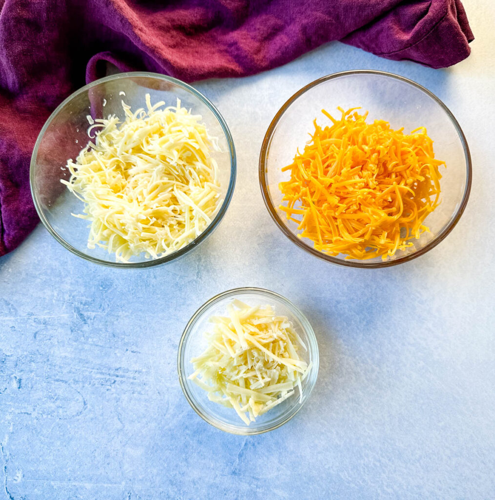 grated cheddar, gruyere, and Parmesan cheese in separate glass bowls