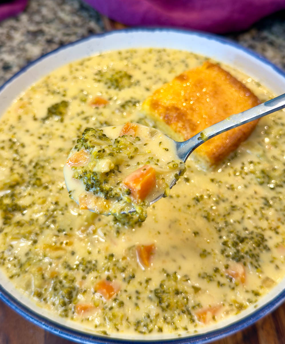 Close-up of a spoonful of broccoli cheddar soup lifted from a bowl, showing tender broccoli and carrots in creamy cheese broth with a square of cornbread in the background.