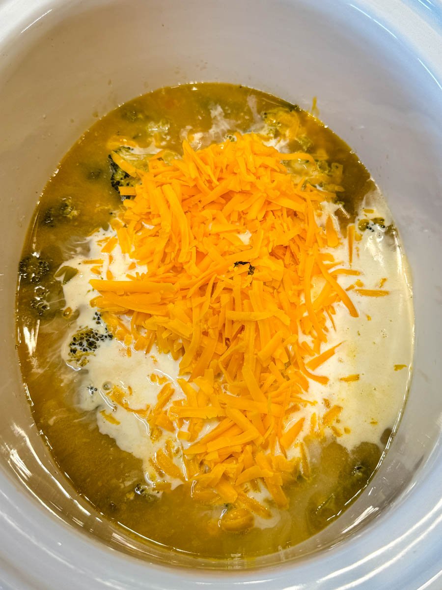 Cheddar cheese melting into a slow cooker filled with broccoli soup and cream, creating a rich cheesy base.