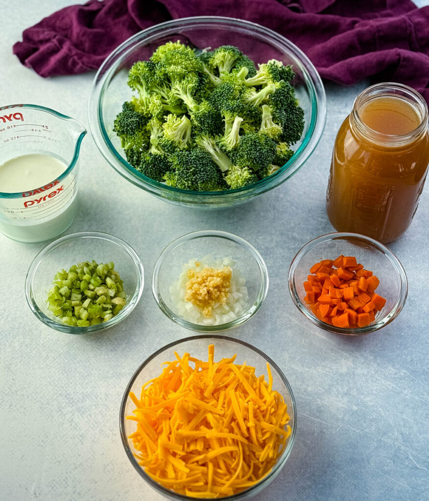 Glass bowls filled with ingredients for broccoli cheddar soup including fresh broccoli florets, shredded cheddar cheese, diced carrots, celery, onions with garlic, a measuring cup of cream, and a jar of broth.