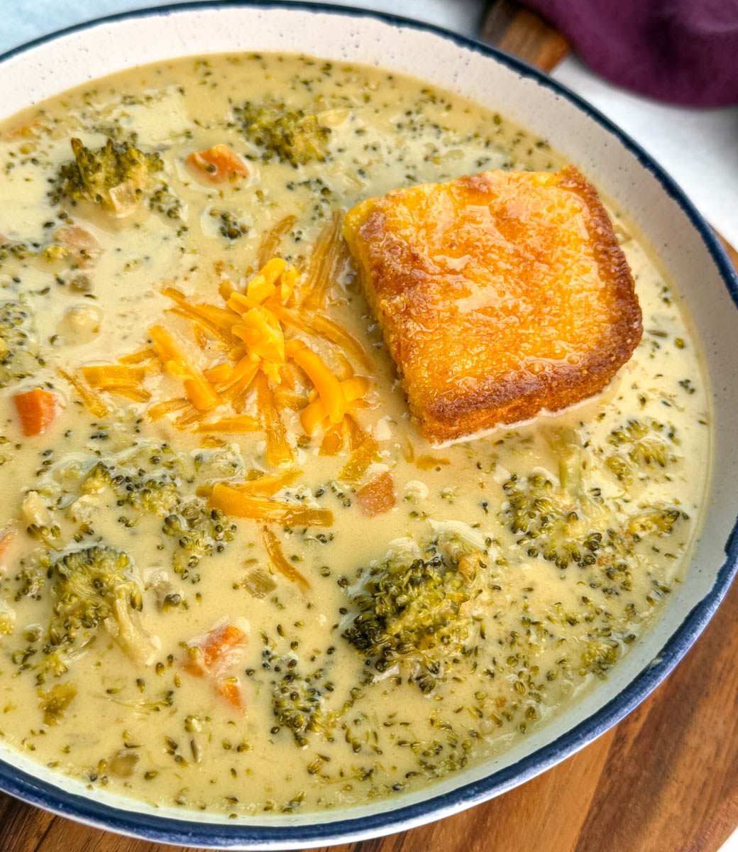Bowl of broccoli cheddar soup topped with shredded cheddar cheese and served with a square of golden cornbread on the side.