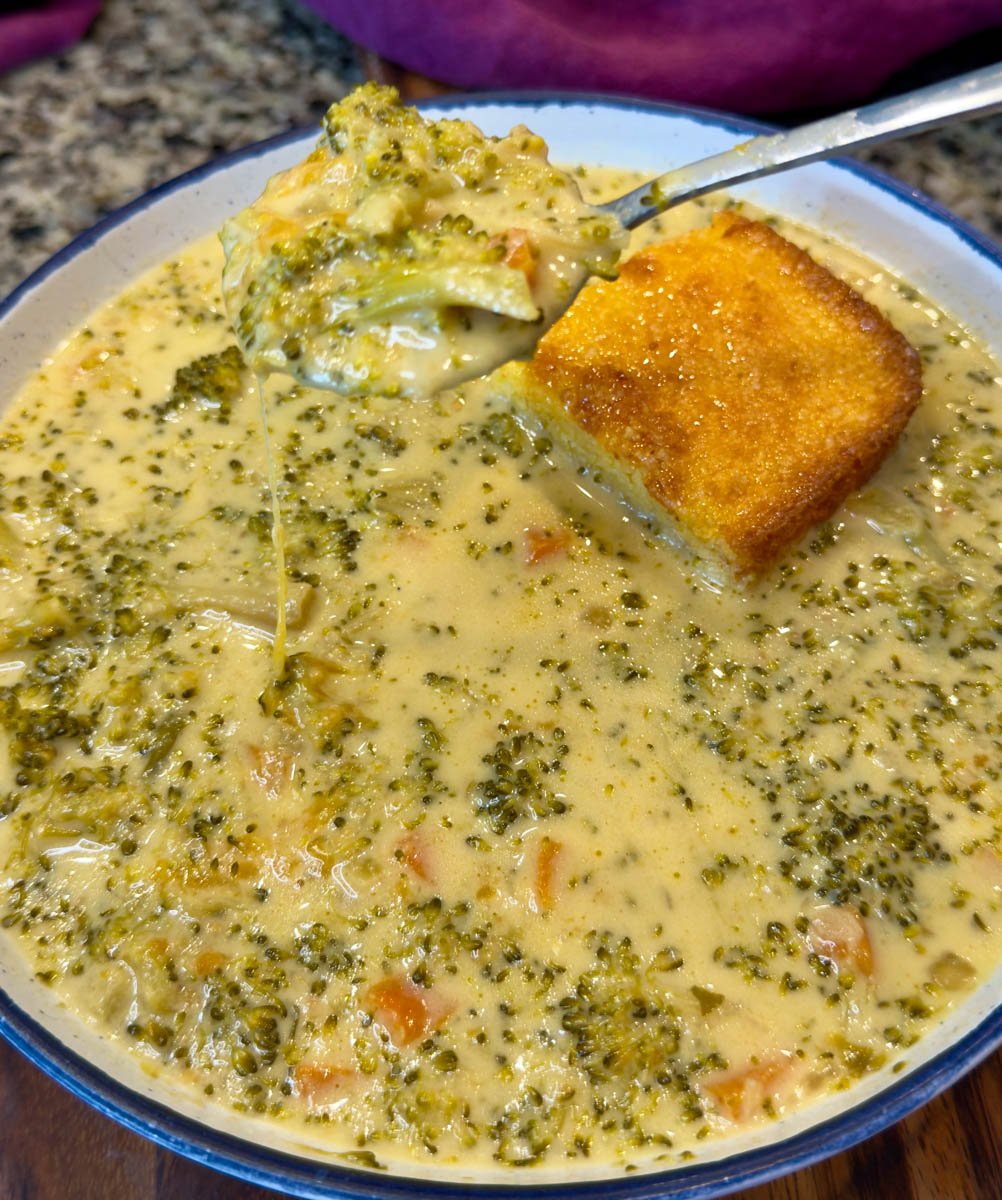 Close-up of a spoonful of cheesy broccoli cheddar soup lifted from a bowl, showing melted cheese strands, tender broccoli, and carrots, with a golden square of cornbread in the soup.