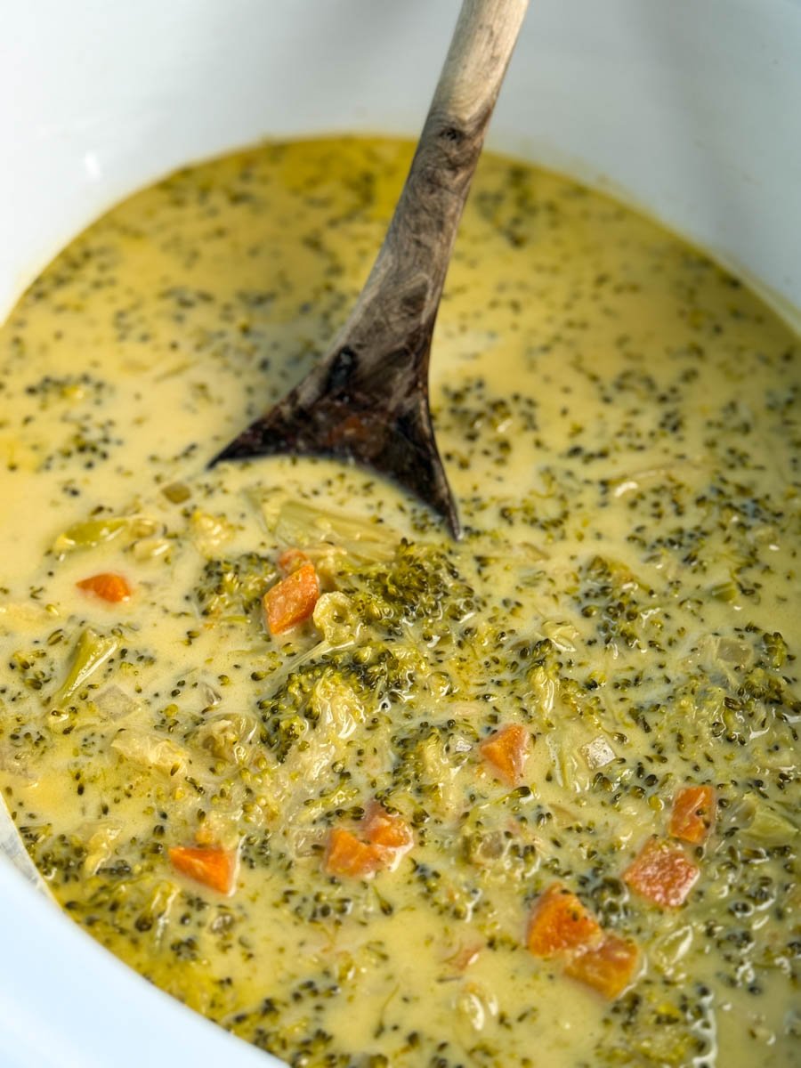 Close-up of broccoli cheddar soup in a slow cooker, with visible pieces of broccoli and carrots in a creamy cheese broth.