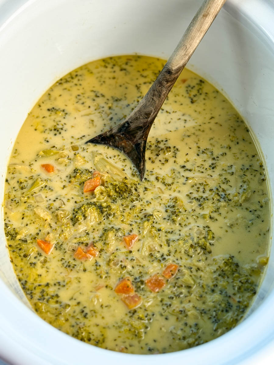 Slow cooker filled with creamy broccoli cheddar soup, showing chunks of broccoli and carrots, with a wooden spoon resting inside.