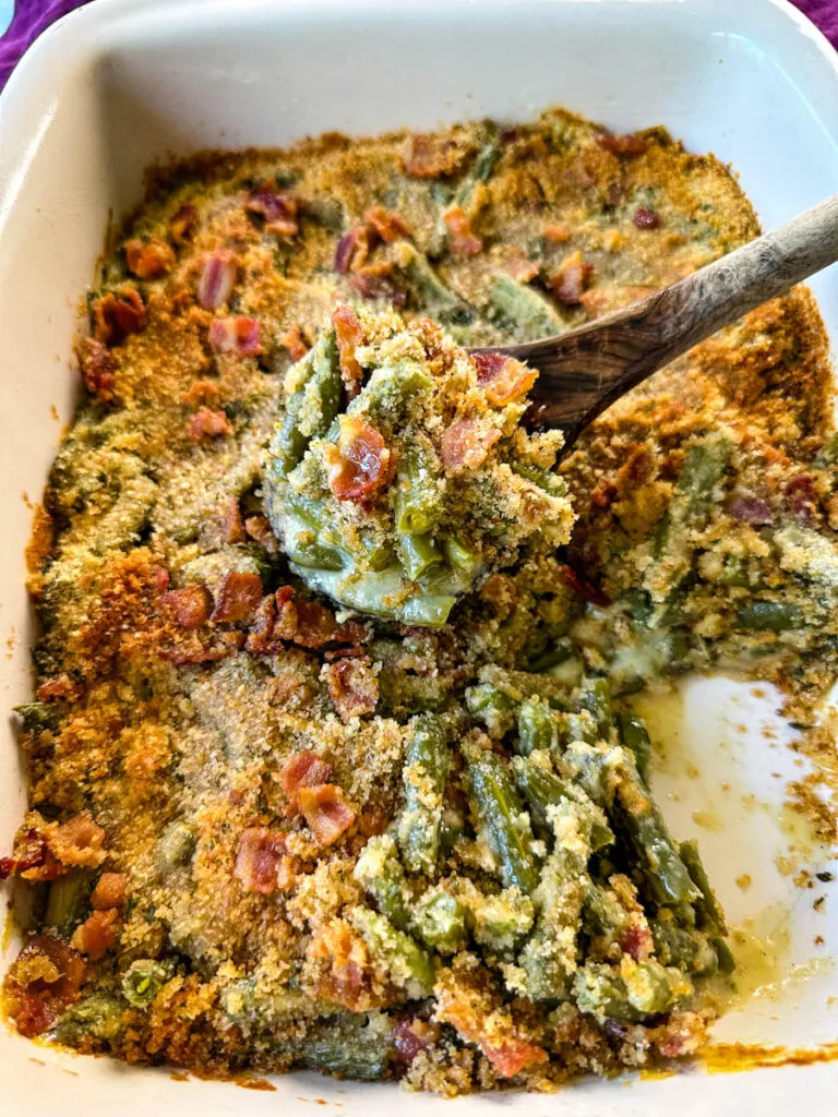 a wooden spoonful of green bean casserole with bacon and breadcrumbs in a baking dish