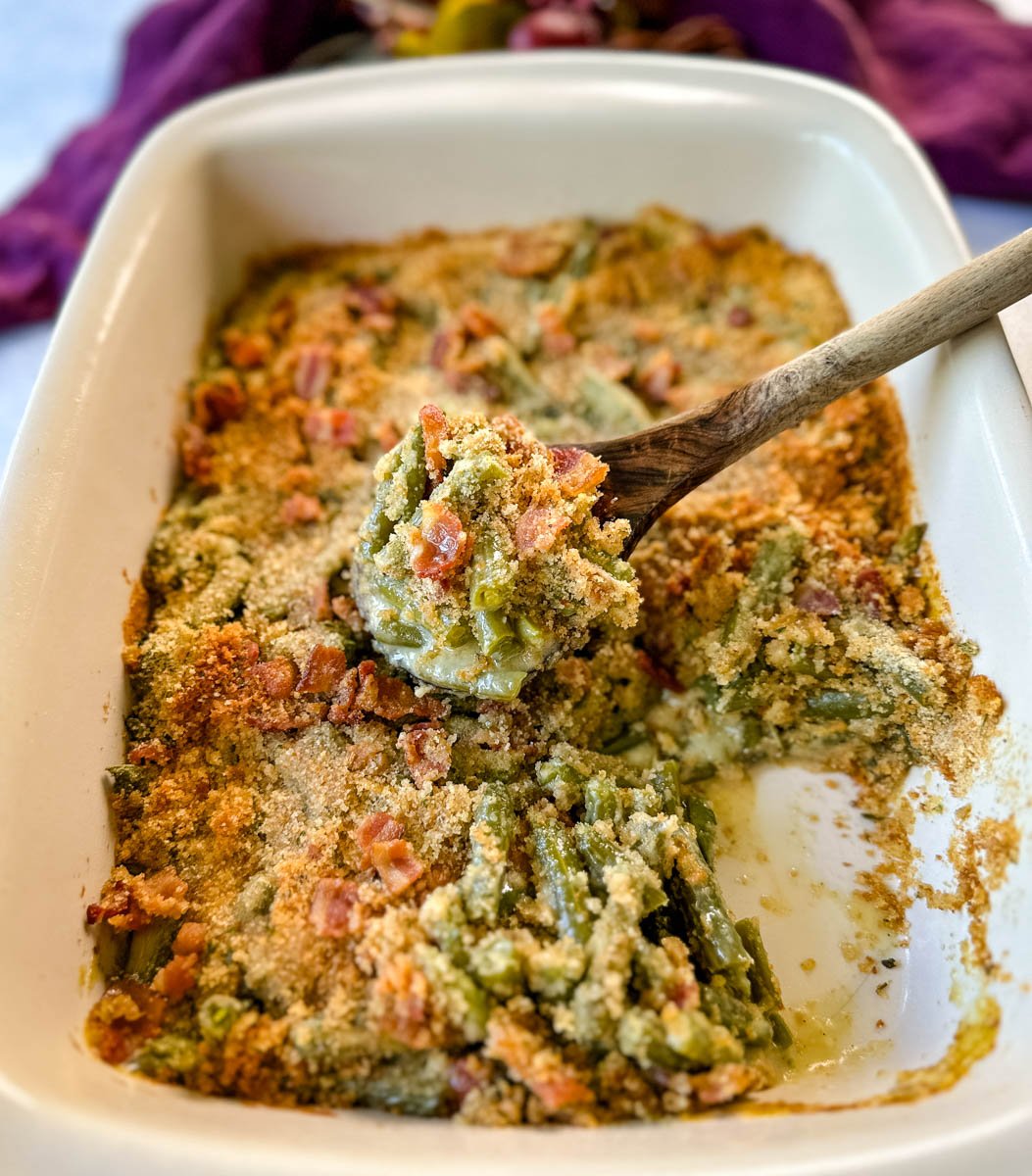 a wooden spoonful of green bean casserole with bacon and breadcrumbs in a baking dish