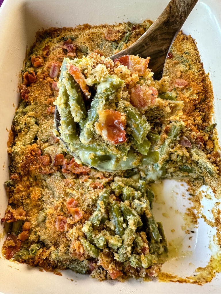 a wooden spoonful of green bean casserole with bacon and breadcrumbs in a baking dish