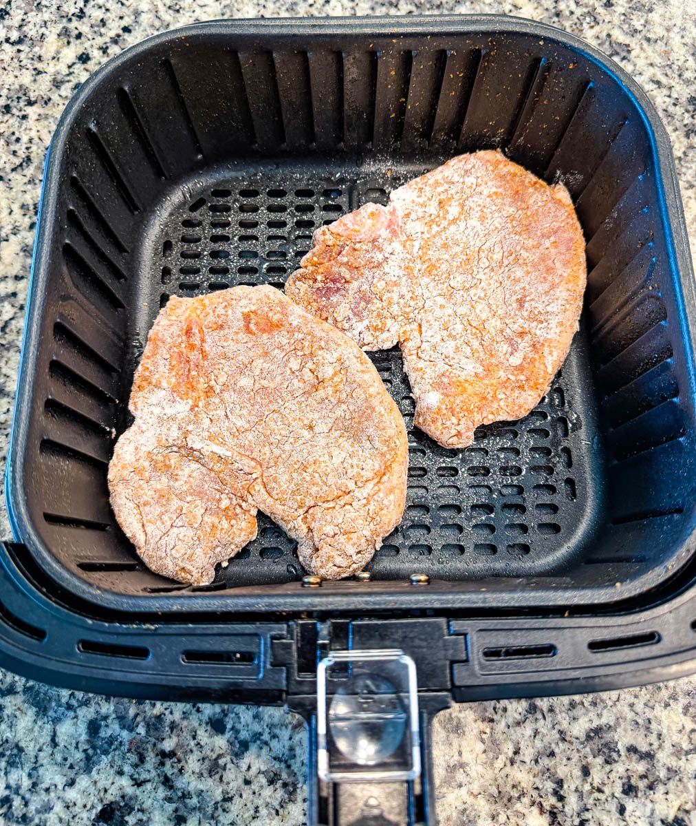 Two raw, breaded pork chops placed in the air fryer basket before cooking.