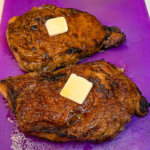Air fryer ribeye steaks with a deeply browned, seasoned crust resting on a purple cutting board, topped with melting pats of butter.