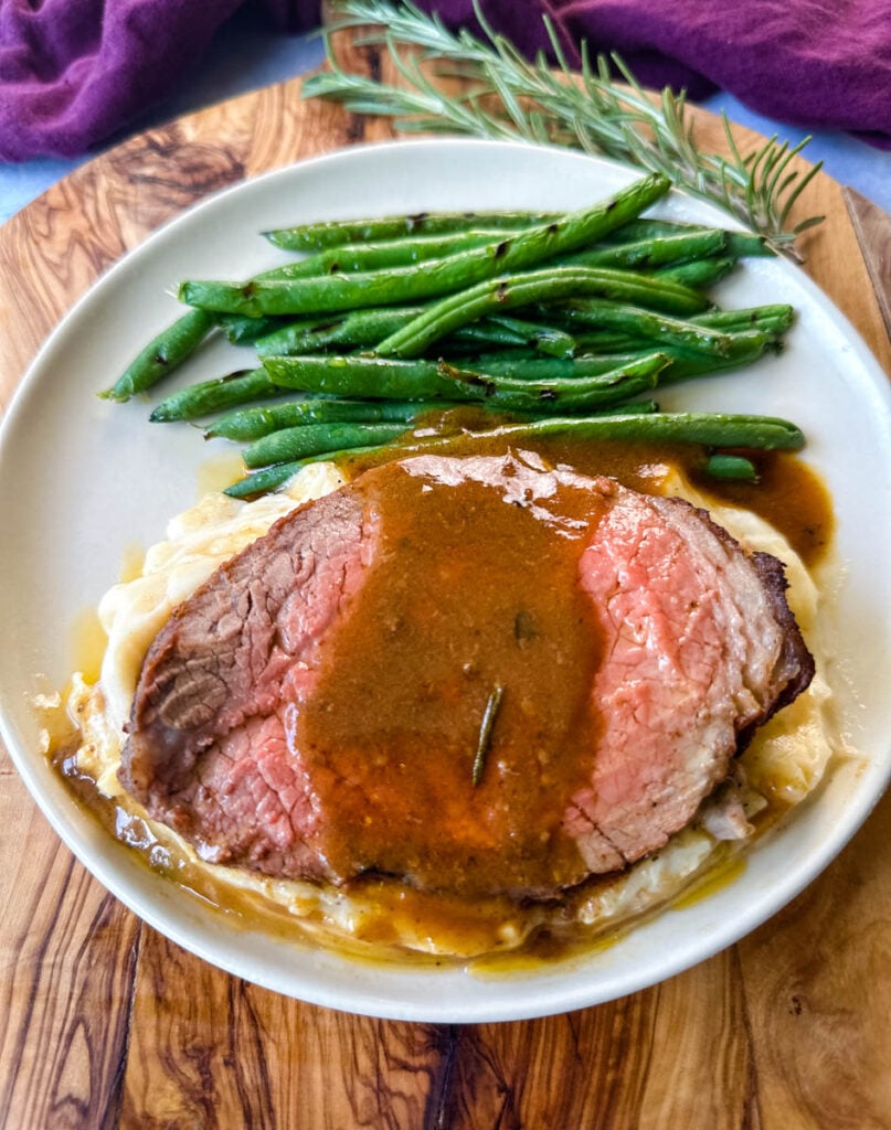 sliced eye of round roast beef on a plate with mashed potatoes, green beans, and au jus gravy