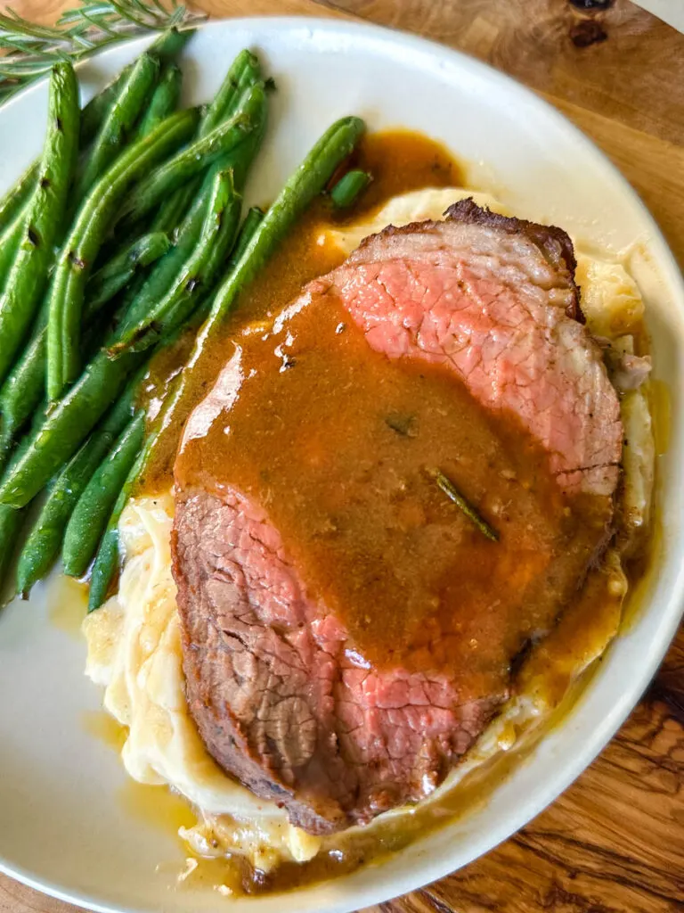 sliced eye of round roast beef on a plate with mashed potatoes, green beans, and au jus gravy