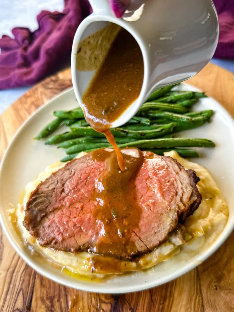 sliced eye of round roast beef on a plate with mashed potatoes, green beans, and drizzled with au jus gravy
