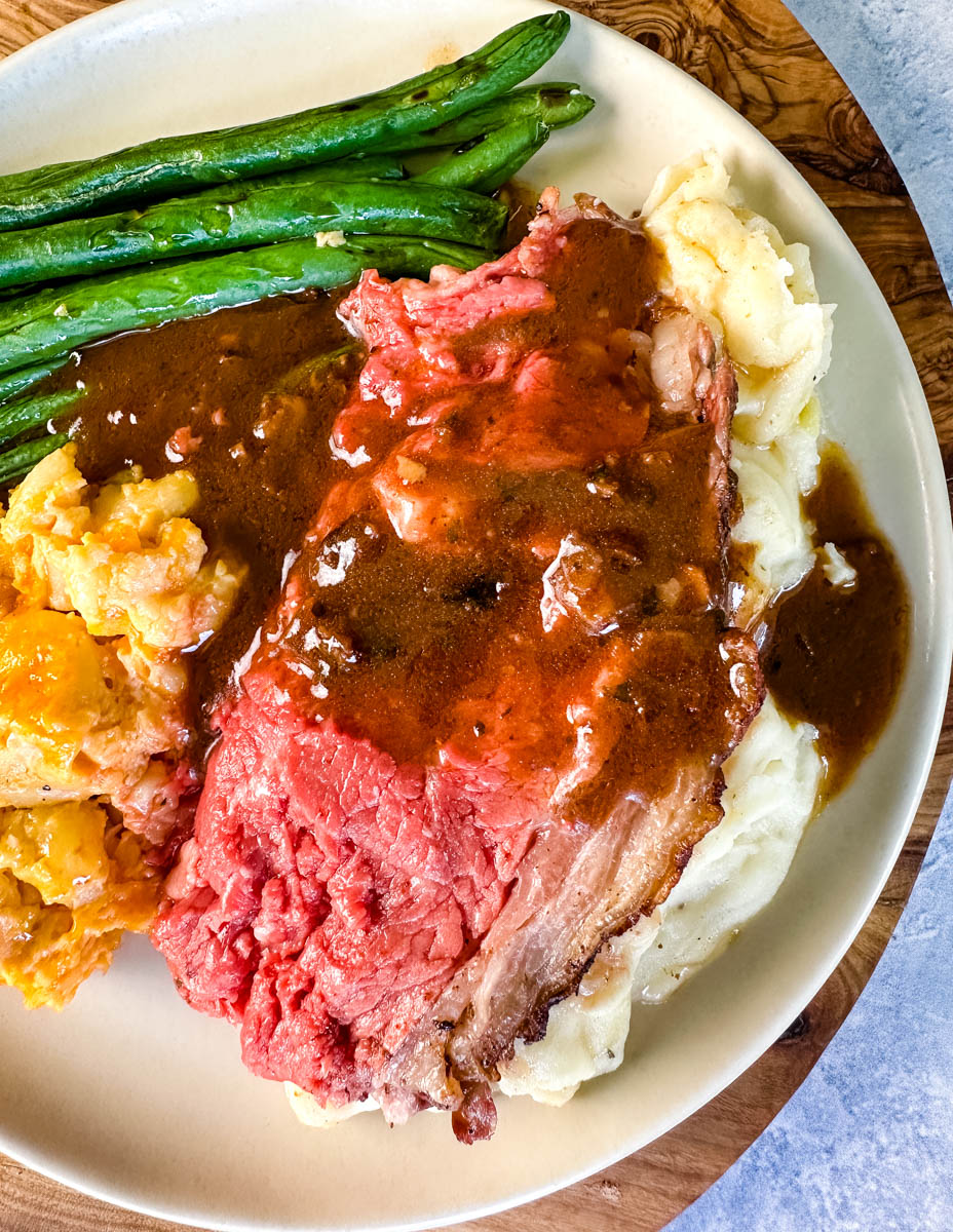 garlic herb butter roasted prime rib with au jus gravy on a plate with green beans, mashed potatoes, and mac and cheese