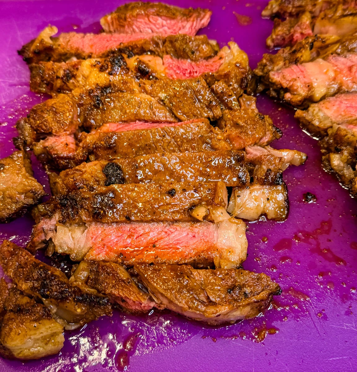 Sliced medium-rare ribeye steak on a purple cutting board, revealing a warm pink center and crispy seasoned edges.