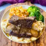 braised beef short ribs drizzled in gravy with mashed potatoes, mac and cheese, and broccoli on a plate