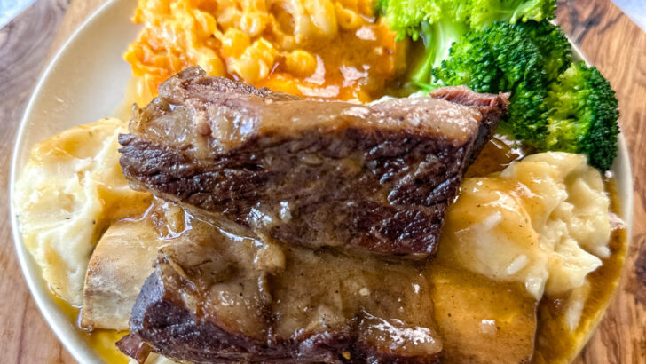 braised beef short ribs drizzled in gravy with mashed potatoes, mac and cheese, and broccoli on a plate