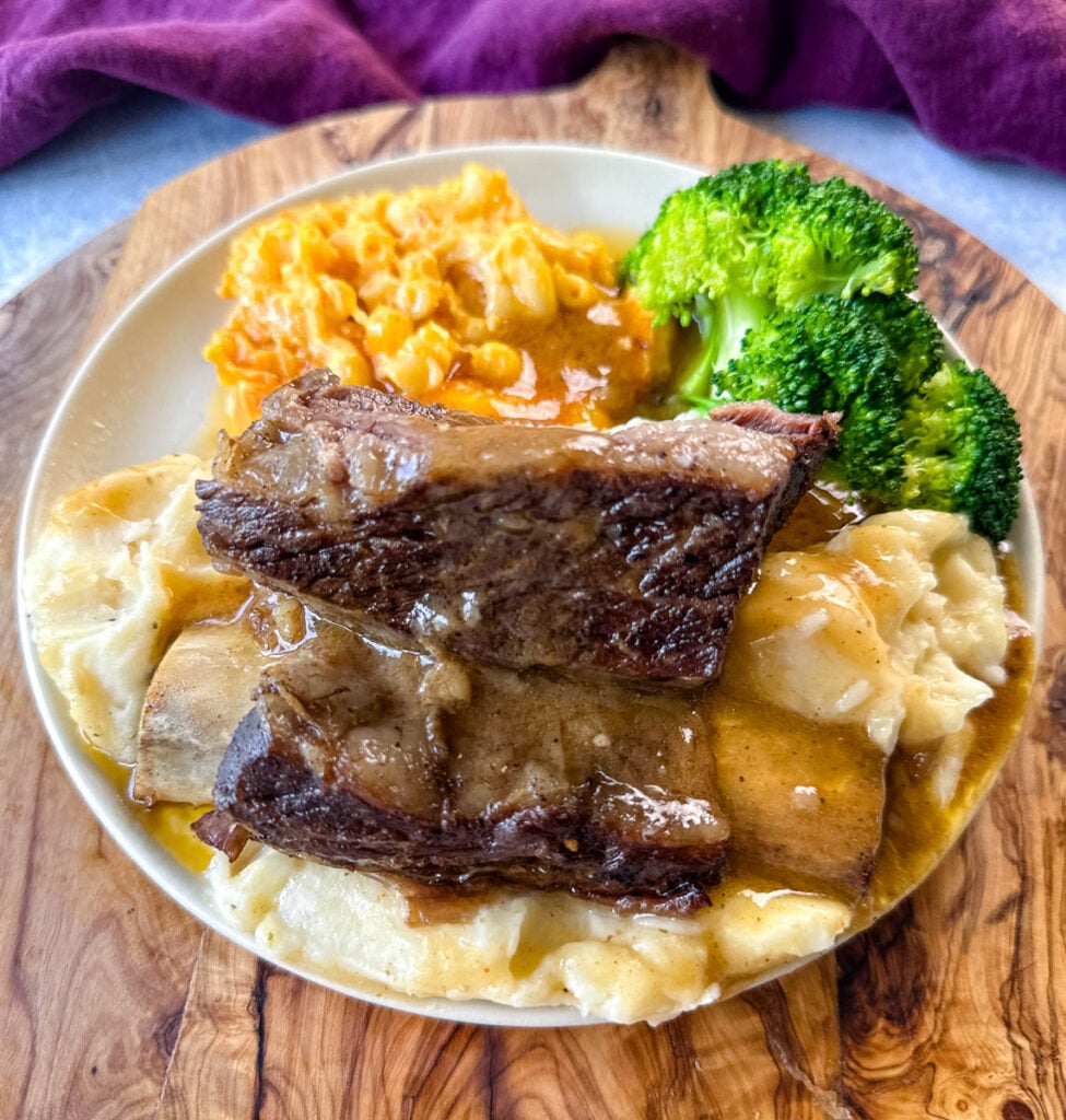 braised beef short ribs drizzled in gravy with mashed potatoes, mac and cheese, and broccoli on a plate