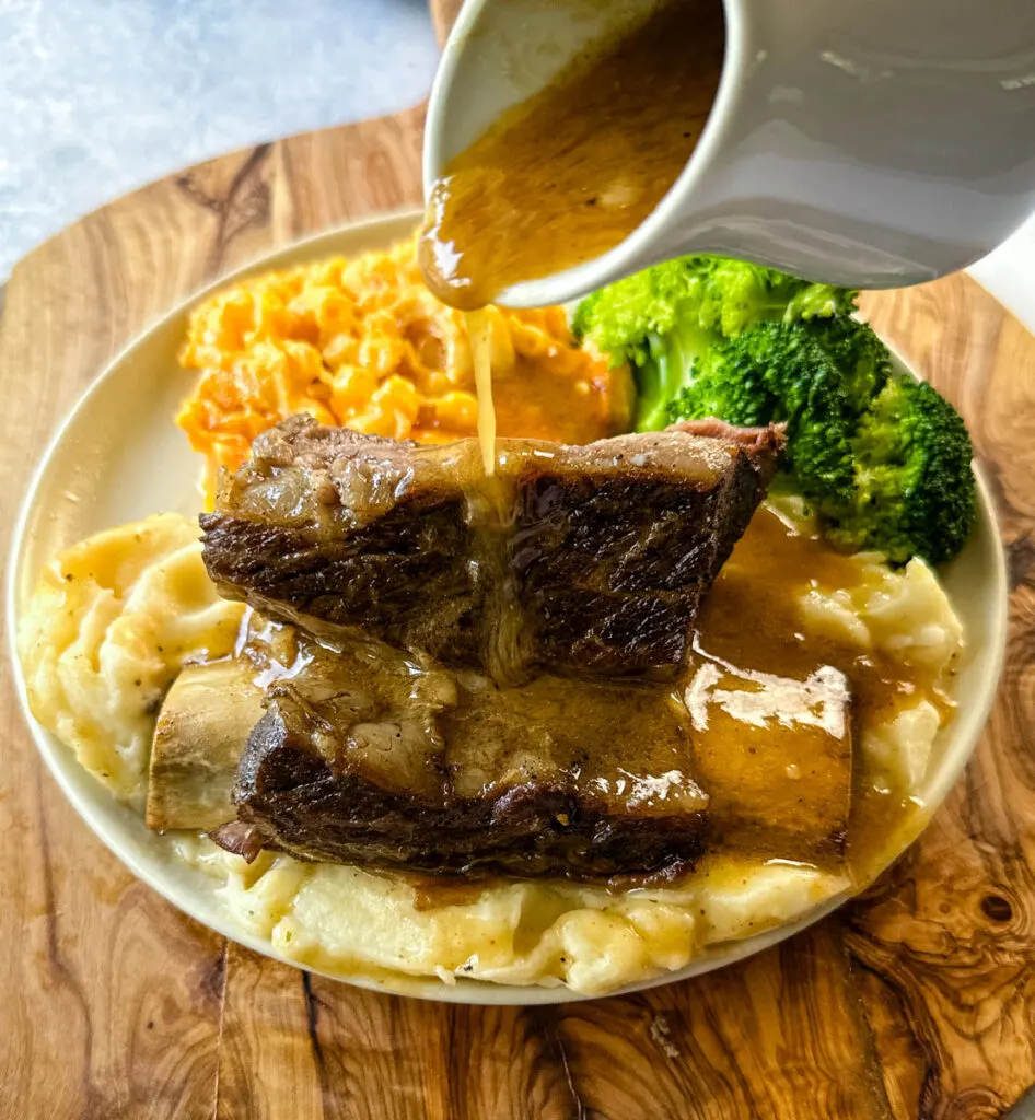 braised beef short ribs drizzled in gravy with mashed potatoes, mac and cheese, and broccoli on a plate