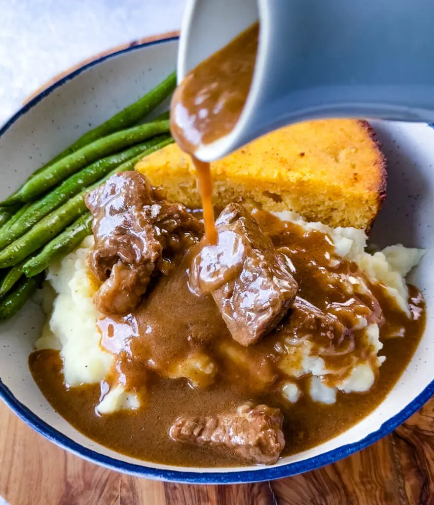 gravy drizzled over beef tips, green beans, cornbread, and mashed potatoes in a bowl