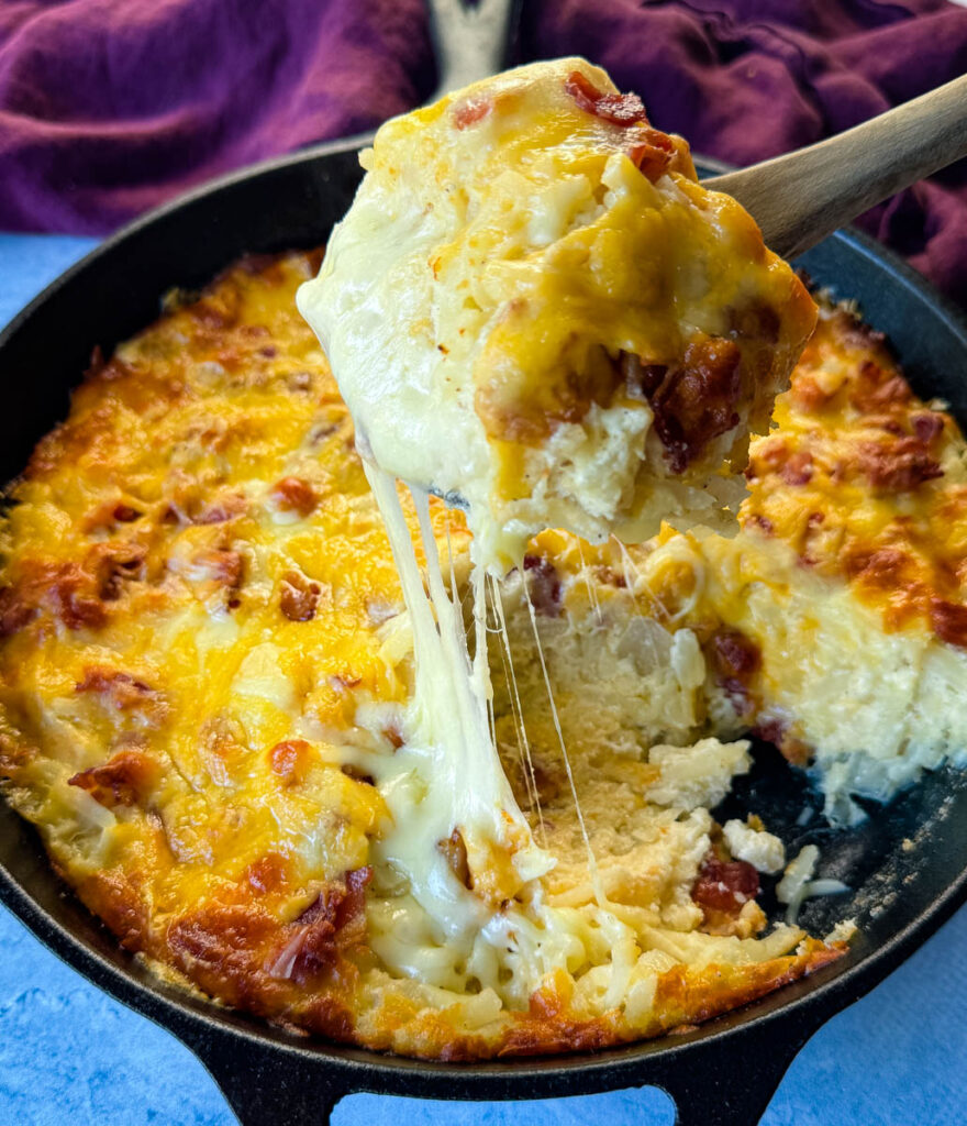 a wooden spoonful of hashbrown casserole with bacon and cheese in a cast iron skillet