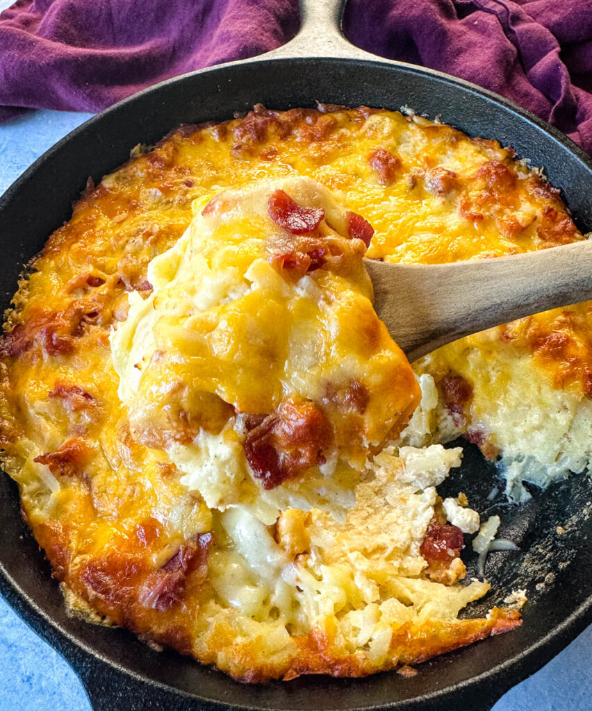 a wooden spoonful of hashbrown casserole with bacon and cheese in a cast iron skillet