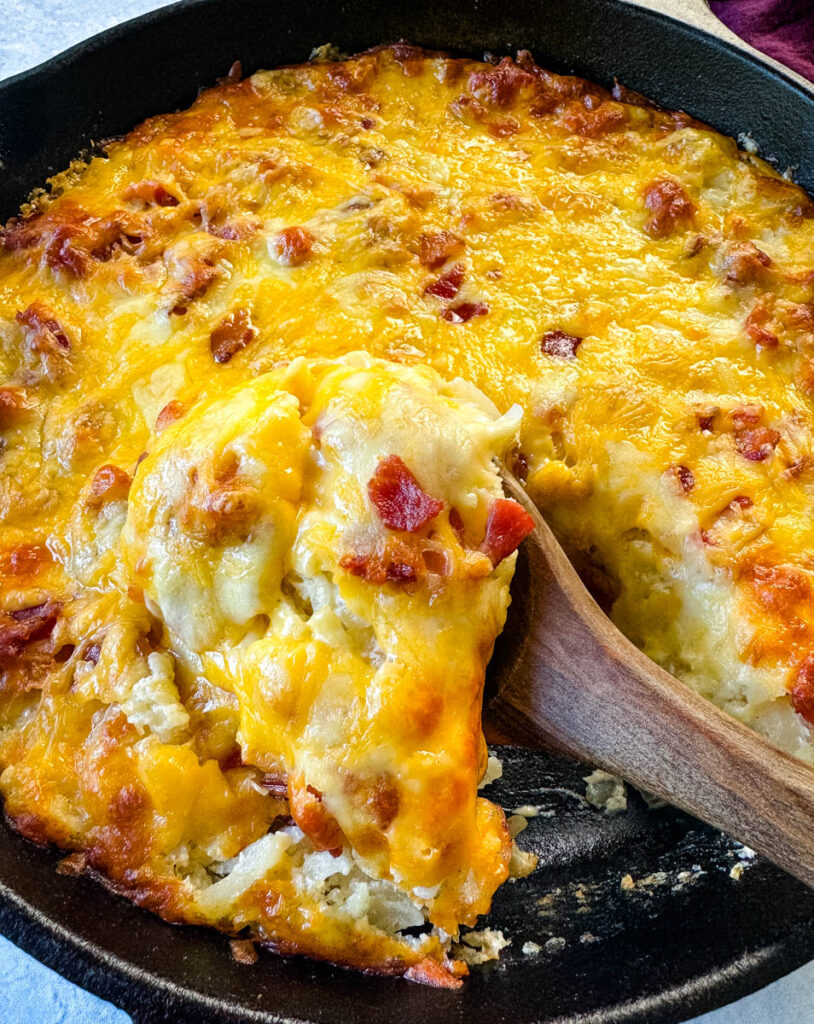 a wooden spoonful of hashbrown casserole with bacon and cheese in a cast iron skillet