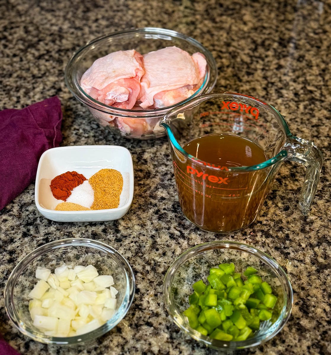 Ingredients for smothered chicken laid out on a countertop including raw chicken thighs in a glass bowl, seasonings, diced onions, diced green bell peppers, and a measuring cup of broth.