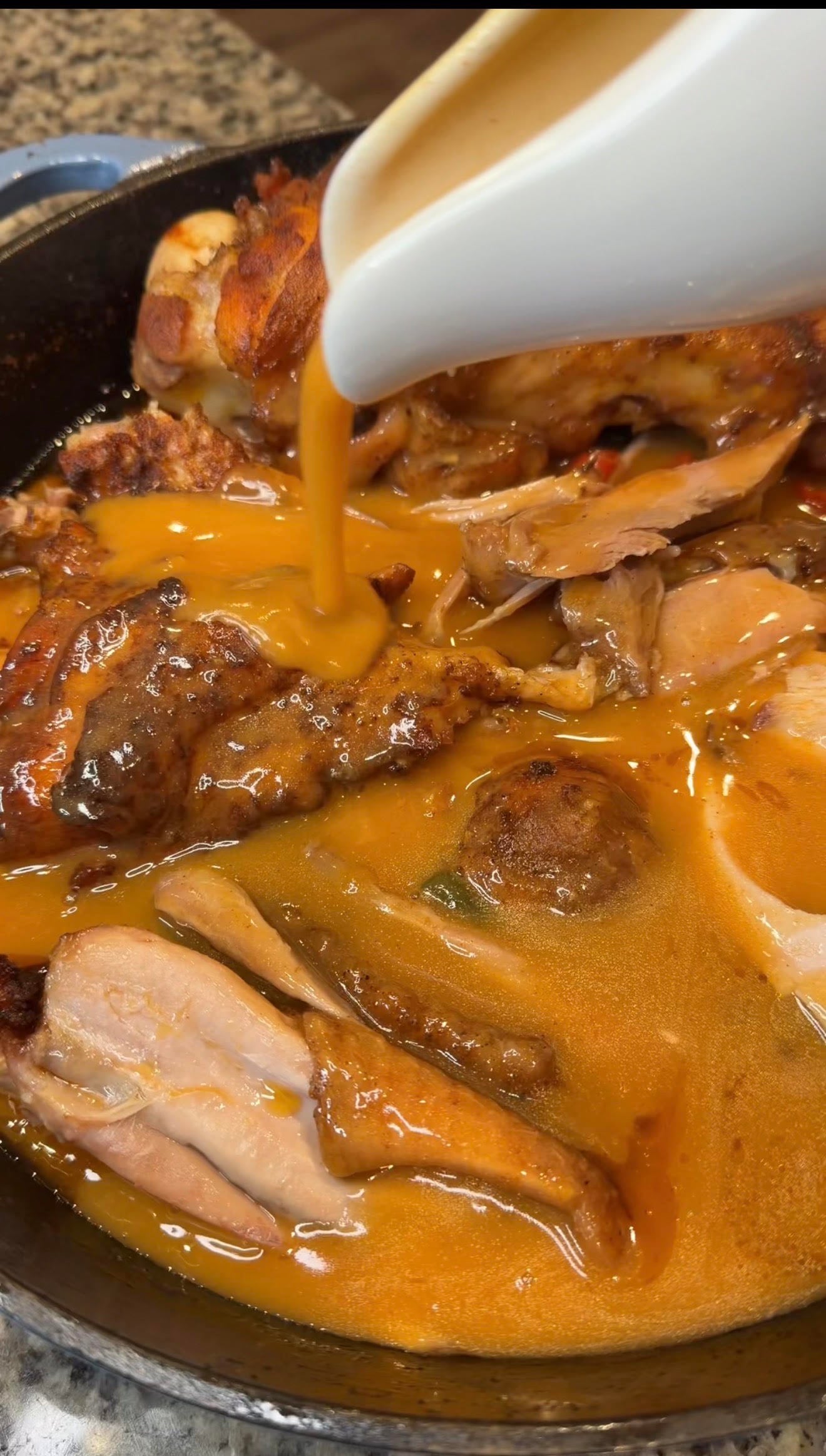 Golden turkey gravy is poured from a white gravy boat over roasted turkey pieces in a cast iron skillet.