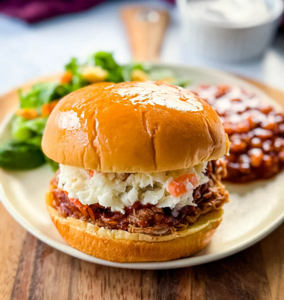 slow cooker Crockpot pulled chicken sandwich with BBQ sauce and coleslaw on a plate with baked beans