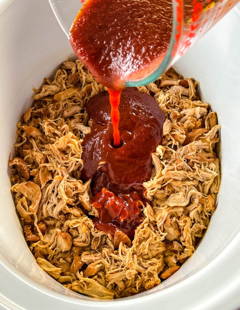 BBQ sauce drizzled over pulled chicken in a white Crockpot slow cooker