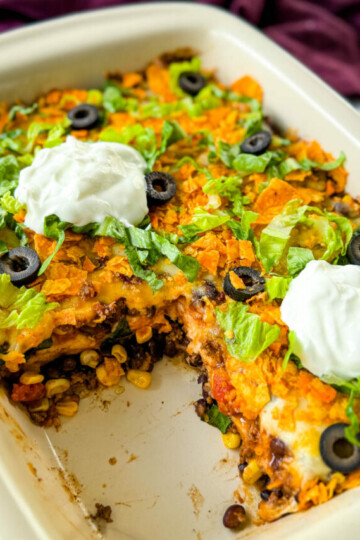 taco bake caserole in a baking dish with Doritos, olives, lettuce, and sour cream