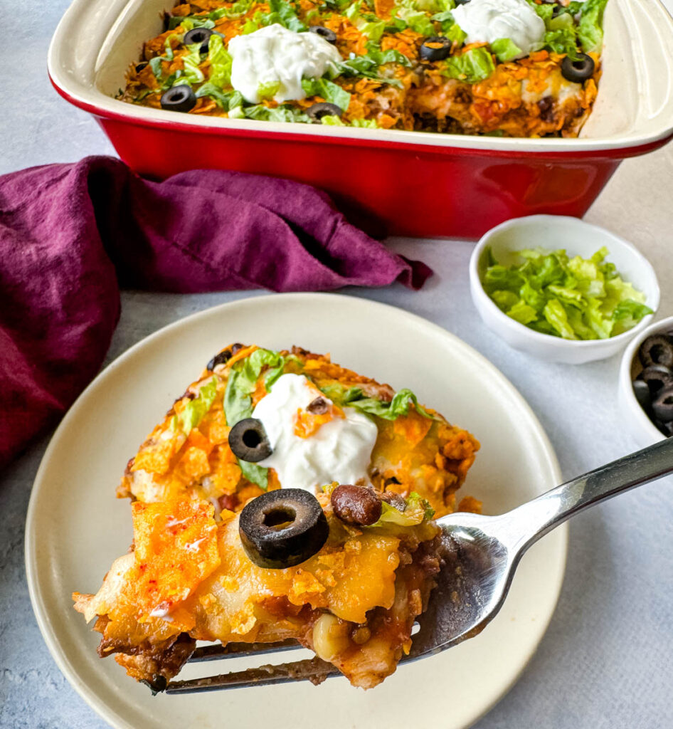 a forkful of taco bake caserole on a plate with Doritos, olives, lettuce, and sour cream