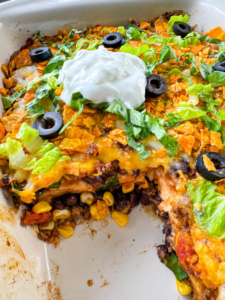 taco bake caserole in a baking dish with Doritos, olives, lettuce, and sour cream