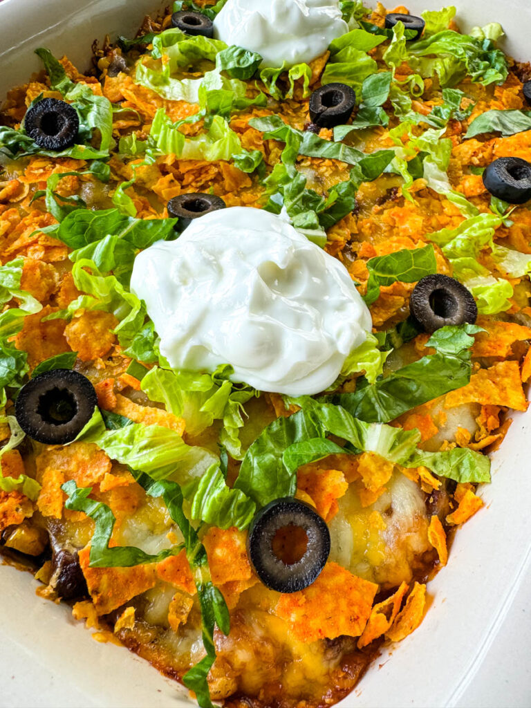 taco bake caserole in a baking dish with Doritos, olives, lettuce, and sour cream