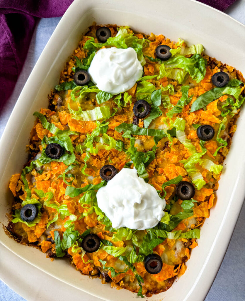 taco bake caserole in a baking dish with Doritos, olives, lettuce, and sour cream