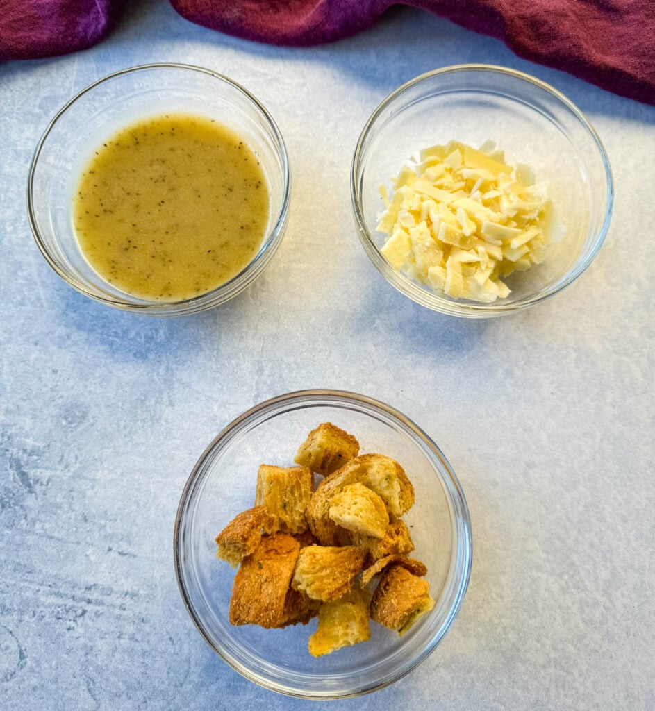 Caesar salad dressing, grated Parmesan cheese, and croutons in separate glass bowls