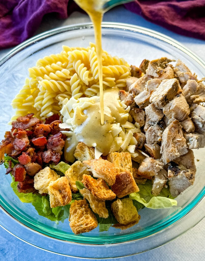 salad dressing drizzled over Chicken Caesar Pasta Salad with bacon and croutons in a glass bowl