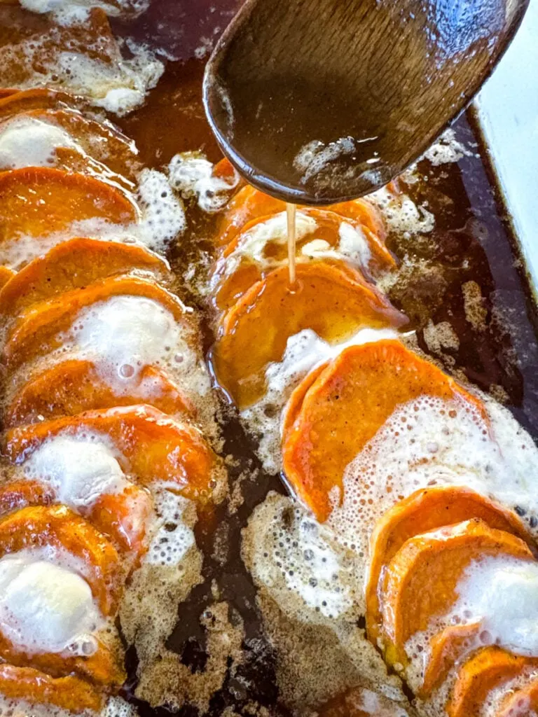 candied sweet potatoes with marshmallows in a baking dish drizzled with glaze in a wooden spoon