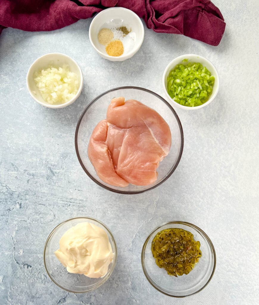 raw chicken breasts, mayo, celery, relish, onions, and spices in separate white bowls