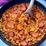 homemade pork and beans with sausage, bacon, and a wooden spoon in a cast iron skillet