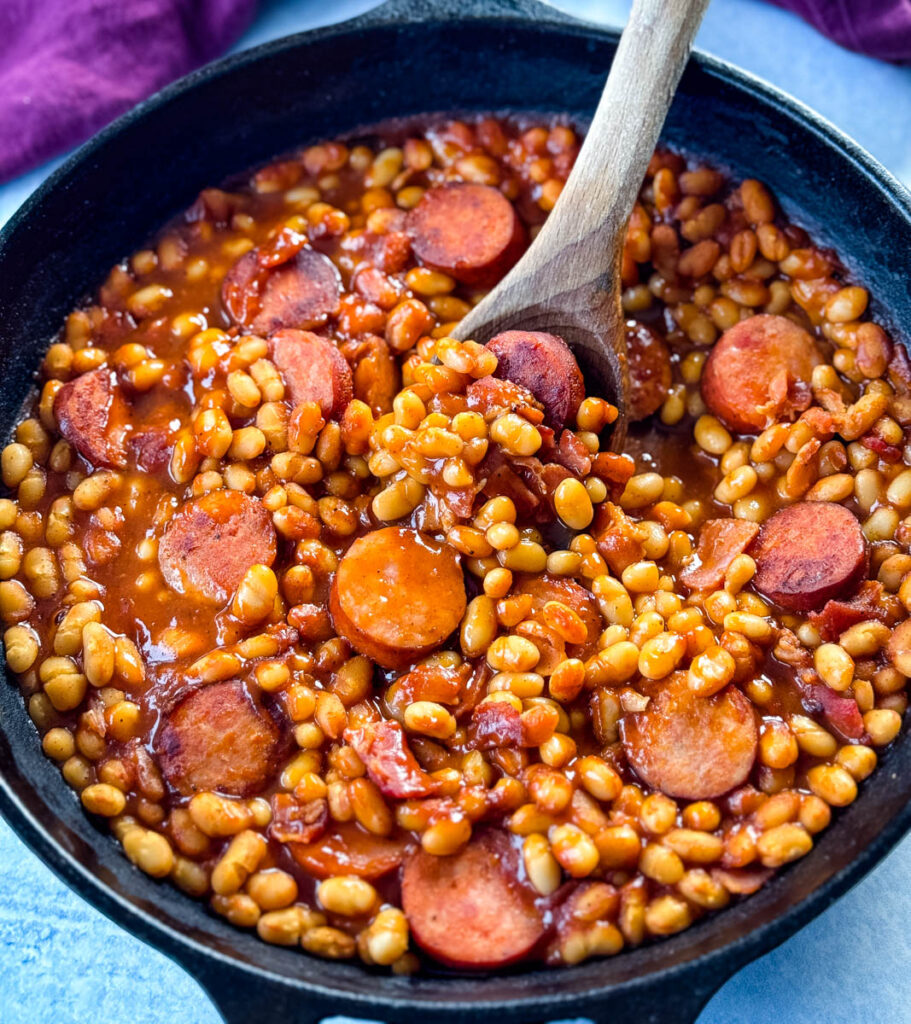 homemade pork and beans with sausage, bacon, and a wooden spoon in a cast iron skillet