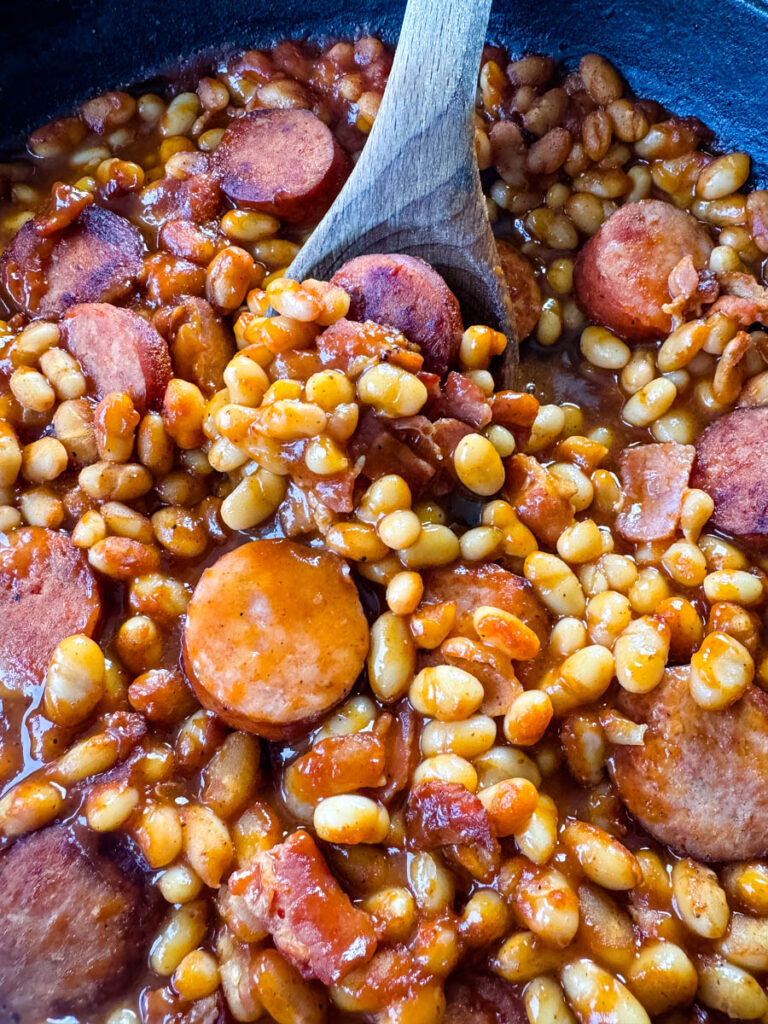 homemade pork and beans with sausage, bacon, and a wooden spoon in a cast iron skillet