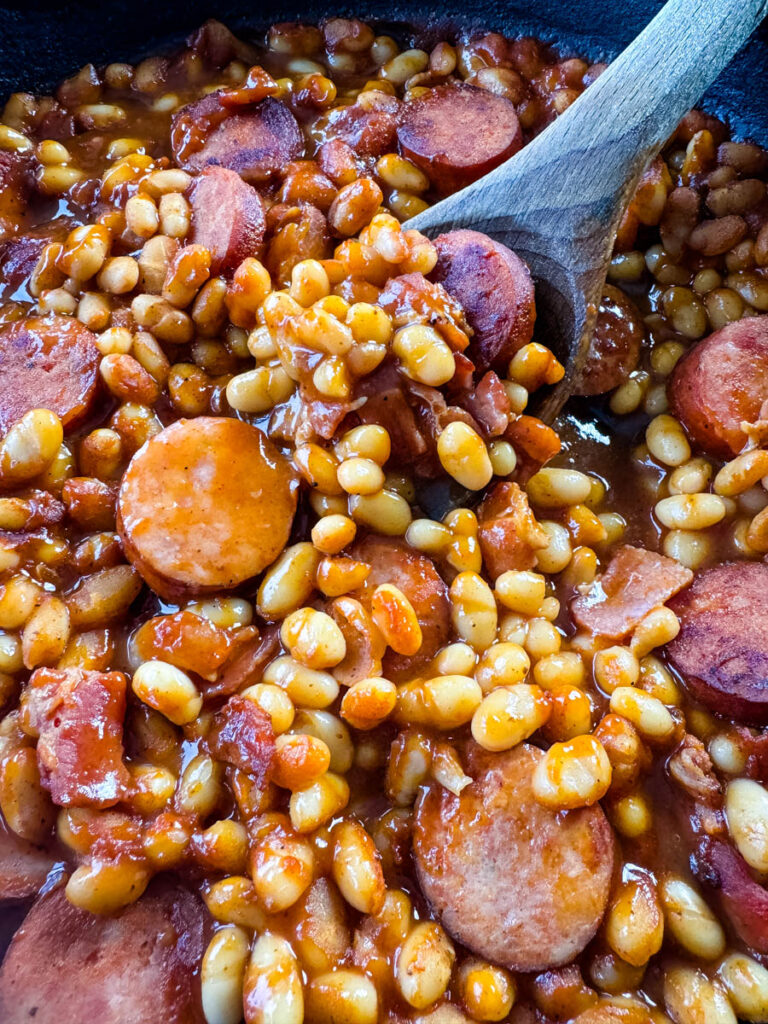 homemade pork and beans with sausage, bacon, and a wooden spoon in a cast iron skillet