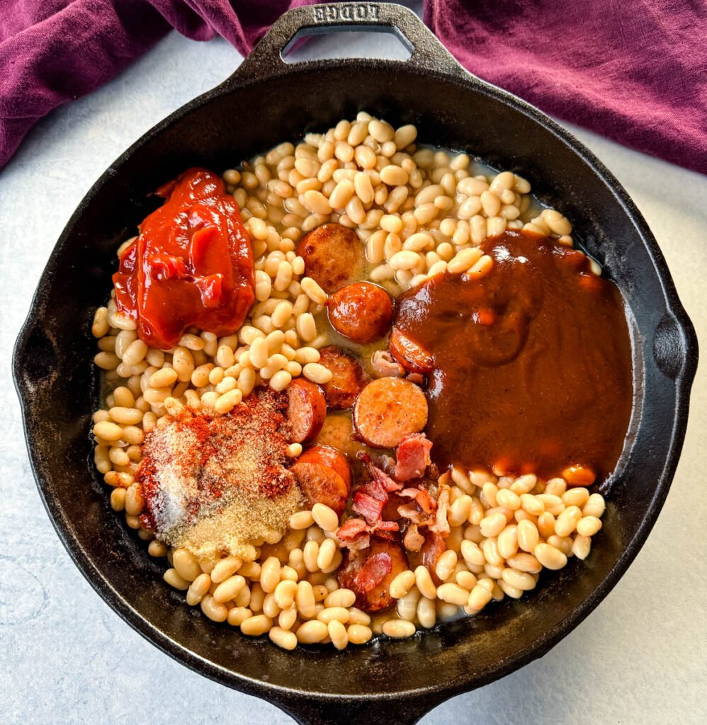 kidney beans with ketchup, BBQ sauce, BBQ rub spices, bacon, and sausage in a cast iron skillet