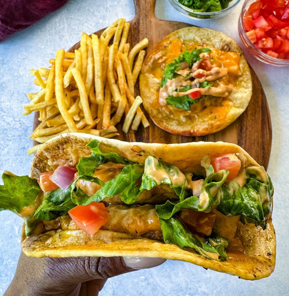 person holding cheeseburger tacos with tomatoes, lettuce, and burger sauce over a plate with fries