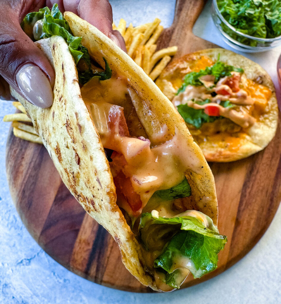 person holding cheeseburger tacos with tomatoes, lettuce, and burger sauce over a plate with fries