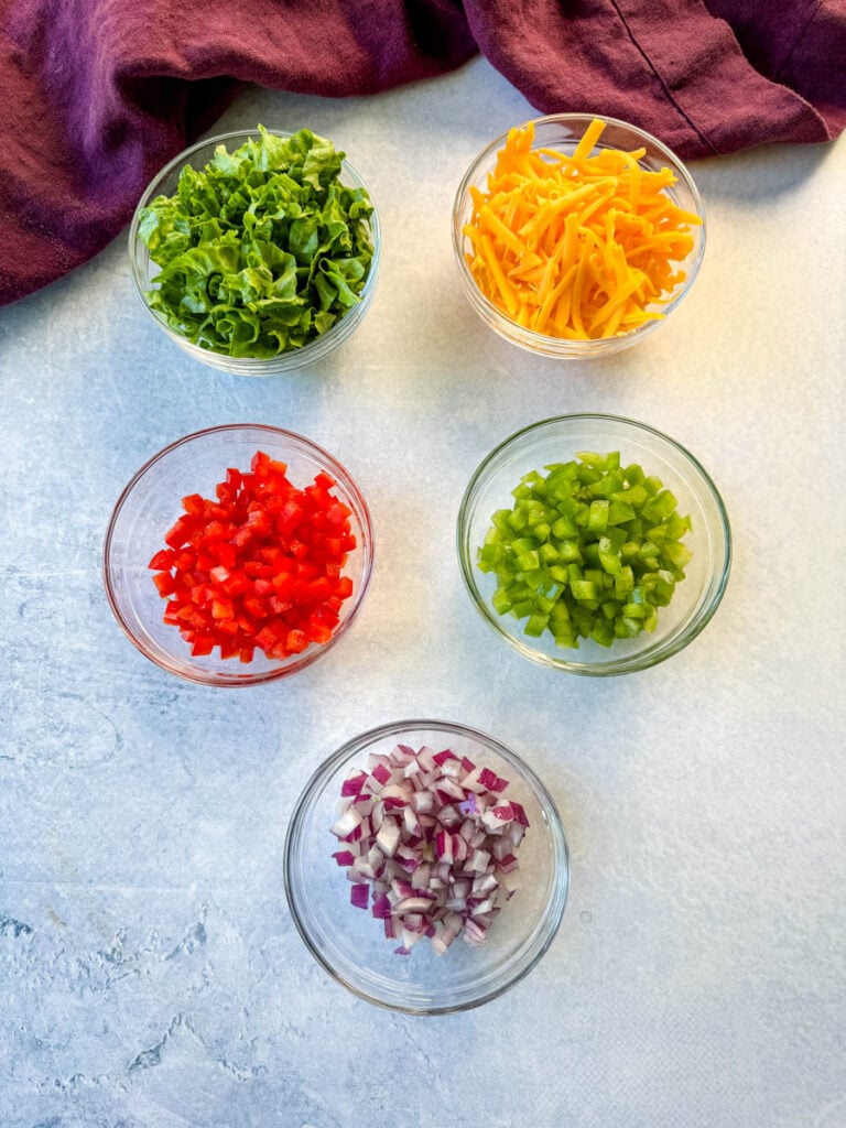 lettuce, grated cheddar cheese, diced red peppers, diced green peppers, and diced onions in separate glass bowls