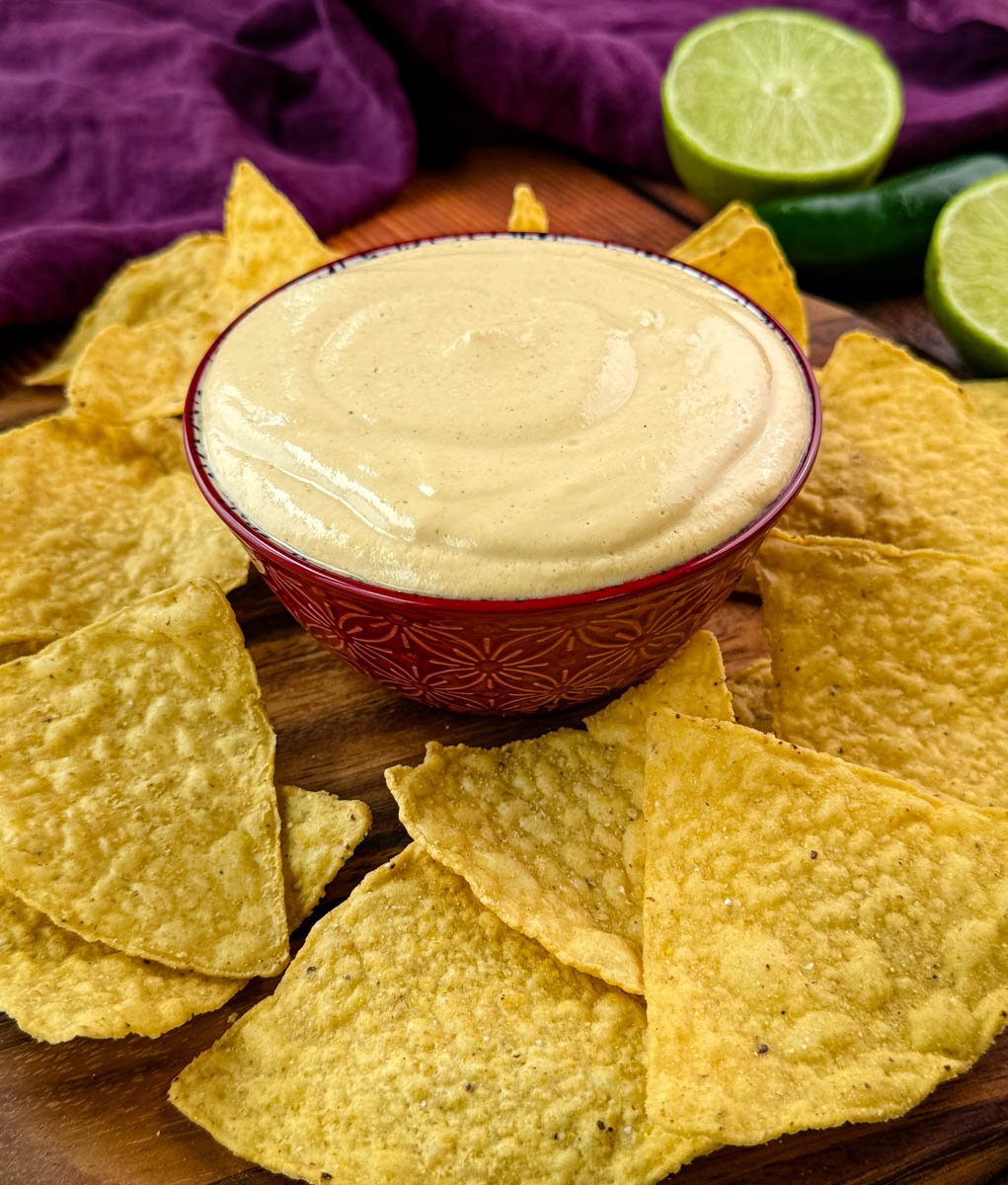 A decorative bowl filled with creamy high-protein queso dip on a wooden board, surrounded by crispy tortilla chips and garnished with lime and jalapeño.