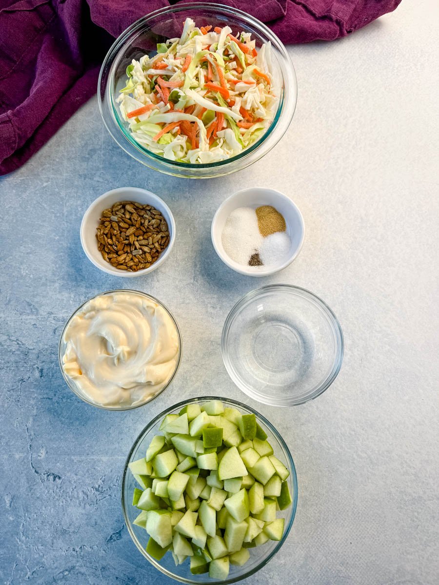 Overhead view of ingredients for creamy apple coleslaw arranged on a light surface, including shredded cabbage and carrots, diced green apples, mayo, seasonings, vinegar, and sunflower seeds.