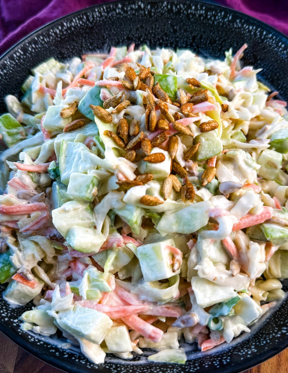 Close-up of creamy apple coleslaw topped with toasted sunflower seeds, showing chopped green apples, shredded cabbage, and carrots in a creamy dressing.